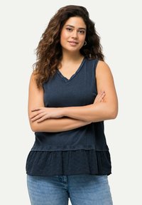 Top sin mangas en azul marino con un dobladillo de peplum texturizado, escote en forma de V y un ligero detalle de volantes, usado sobre unos jeans de un azul claro.