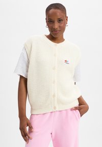 Crèmegebreide vest met een gemêleerd patroon, voorzien van een ronde halslijn, knopen aan de voorkant en een klein, veelkleurig logo op de borst.