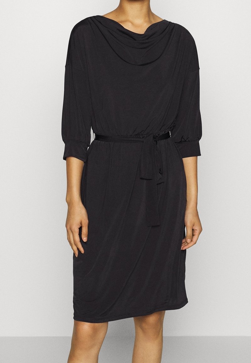 Femme portant une robe noire longueur genou avec des manches trois-quarts, un col bénitier et une ceinture nouée à la taille.