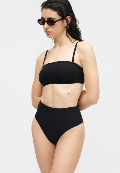 Γυναίκα που φοράει μαύρο μαγιό ψηλόμεσο κατώτερο μέρος και strapless πάνω μέρος, με σκοτεινά γυαλιά ηλίου και βρεγμένα, πίσω χτενισμένα μαλλιά, ποζάροντας μπροστά σε λευκό φόντο.