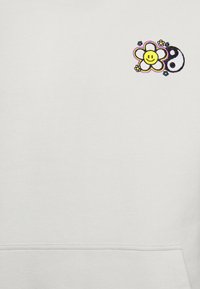 Tissu blanc avec un petit motif brodé d'une fleur jaune souriante et un symbole yin-yang noir et blanc près du coin supérieur droit.