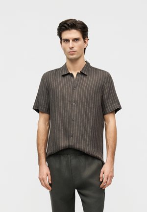 Νεαρός άνδρας που φοράει σκούρο καφέ κοντομάνικο πουκάμισο με λεπτές λευκές κάθετες ρίγες και σκούρο παντελόνι, μπροστά σε απλό φόντο.