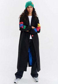 Kobieta nosząca długi czarny płaszcz z kolorowymi literami na rękawach, zieloną czapkę beanie, białe trampki, czarne spodnie, stojąca z założonymi ramionami.