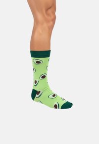 Grüne Socken mit einem sich wiederholenden Avocado-Muster in braunen, schwarzen und weißen Details. Der Bund ist dunkelgrün und der Stoff wirkt weich.