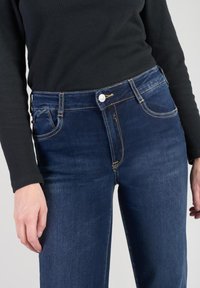 Jeans de un azul oscuro con corte recto, cinco bolsillos, detalles de costura sutiles y cierre de botón plateado. Combinados con una blusa negra de manga larga.