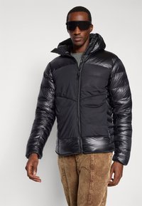 Homme portant une doudoune noire avec capuche, des lunettes de soleil noires, une chemise olive et un pantalon en velours côtelé marron, se tenant devant un fond clair uniforme.