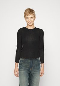Schwarzes, geripptes Langarmshirt mit rundem Halsausschnitt und gewelltem Saum. Getragen mit hellblauen Jeans im lockeren Schnitt.