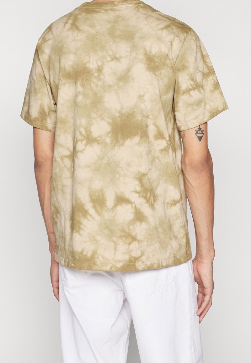 Homme portant une chemise tie-dye beige et marron clair à manches courtes et un pantalon blanc, vu de dos, avec un petit tatouage géométrique sur le bras gauche.