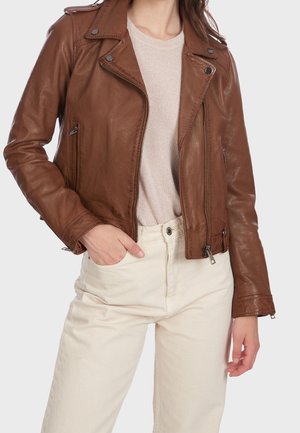 Vrouw draagt een bruine leren bikerjack over een beige trui met lichtgekleurde broek, linkerhand in de zak, rechterarm ontspannen.