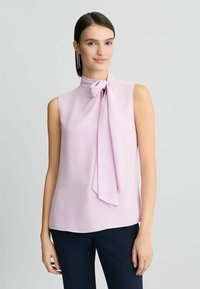 Mouwloze blouse van lichtroze stof met een hoge hals en een strikdetail. Soepele textuur, valt zachtjes, gecombineerd met donkere broek.