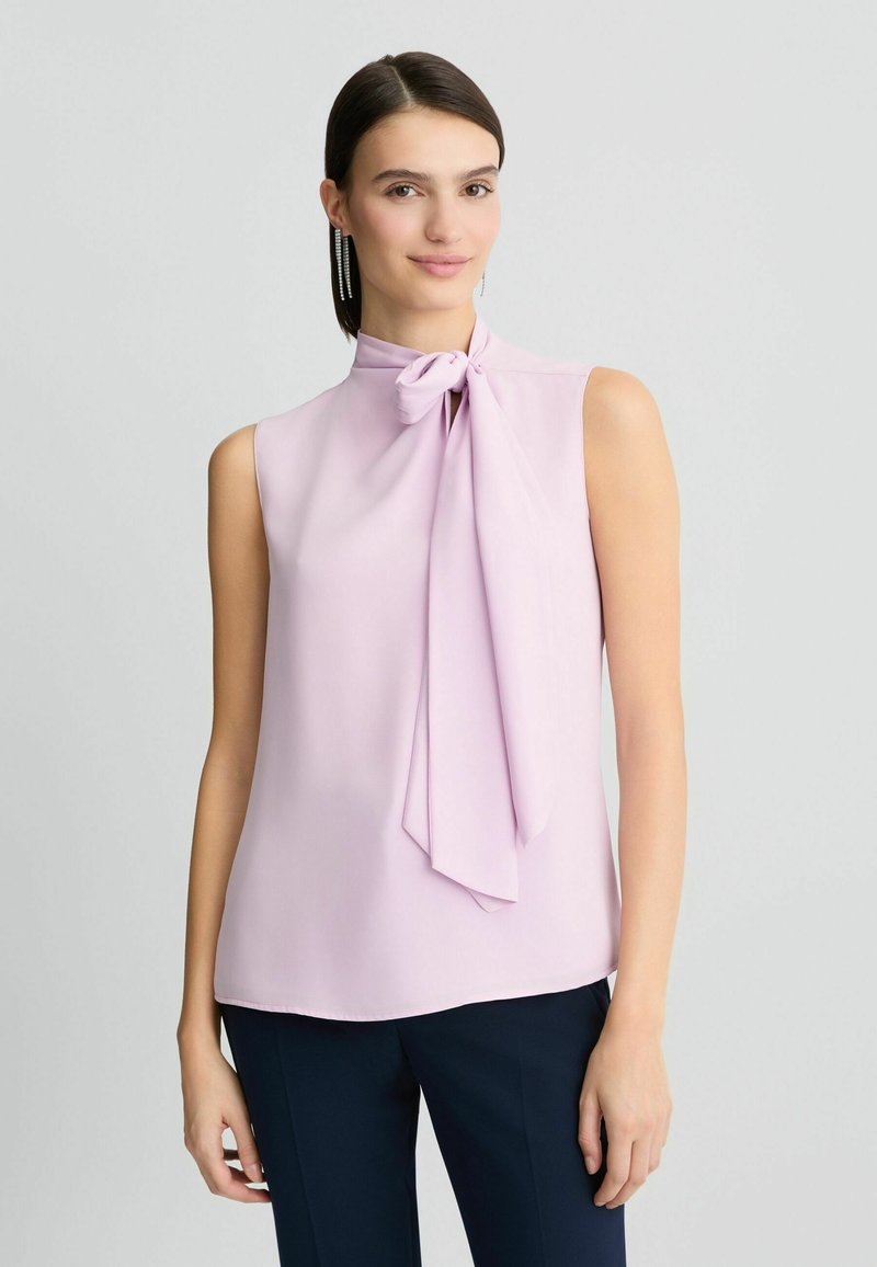 Mouwloze blouse van lichtroze stof met een hoge hals en een strikdetail. Soepele textuur, valt zachtjes, gecombineerd met donkere broek.