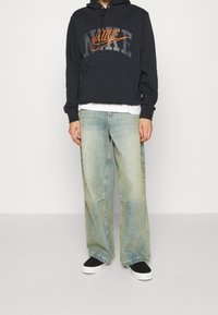 Svart huvtröja med en grå och orange Nike-logotyp, ihop med ljusblå raka jeans och svarta slip-on skor med vita sula.