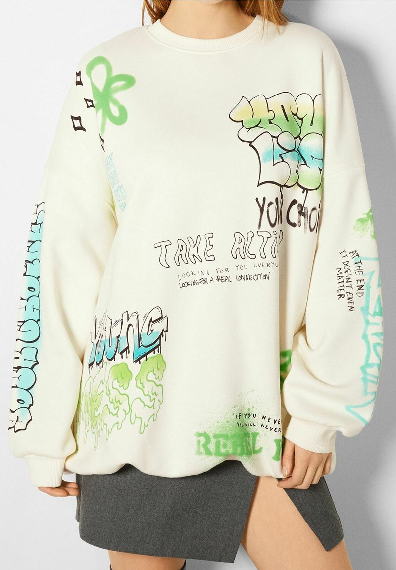 Sudadera extragrande color crema con texto y gráficos de estilo graffiti en verde y azul, con cuello y puños acanalados.