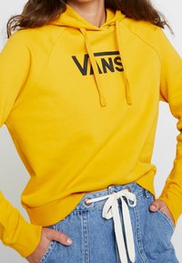 Felpa gialla con logo "VANS" nero, dotata di cappuccio con coulisse e polsini a costine. Abbinata a pantaloni corti in denim a vita alta.