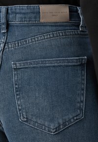Dark blue denim jeans with a back pocket, featuring a leather tag labeled "ANNO 1995 / BLUE DENIM ONLY." Stitched details visible.