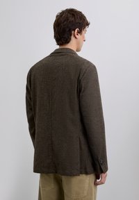 Joven con cabello corto y rizado visto desde atrás, vistiendo un blazer texturizado marrón oscuro y pantalones marrón claro, contra un fondo liso.