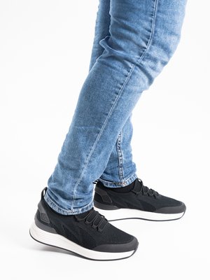 Legs in blue jeans wearing black athletic sneakers with white soles, standing with feet crossed on a white background.