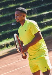 Mees erkor kollases spordirõivastuses, hoides tennisereketit, valmistub mängima välisel savitenniseväljakul, mille taustal on kivist tribüünid.