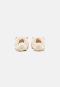 Fuzzy beige slippers with a plush texture, featuring round pom-poms at the heel and small bear ears on the sides. Open back design.