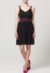 Robe noire avec un col en V et des accents roses aux épaules et à la taille ; tissu fluide, longueur genou, associée à des escarpins roses.