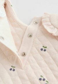 Tessuto trapuntato rosa con piccole ricami a forma di ciliegia. Presenta una chiusura a scatto e un'accente arricciato vicino al colletto. Texture morbida evidente.