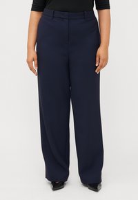 En person bär en åtsittande svart topp och marinblå vida byxor, vilket visar en stilfull outfit kombinerad med svarta högklackade skor mot en vit bakgrund.