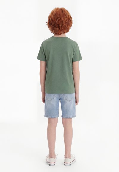 Παιδί με κόκκινα σγουρά μαλλιά στέκεται με την πλάτη, φοράει πράσινο μπλουζάκι, ανοιχτόχρωμα τζιν σορτς και άσπρα αθλητικά παπούτσια σε λευκό φόντο.