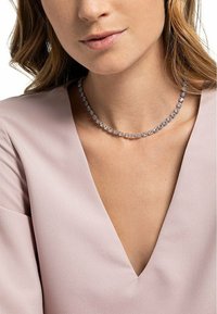 Collana choker in cristallo trasparente con pietre rotonde e sfaccettate montate in metallo tono argento, indossata sopra una blusa a V di colore rosa chiaro.