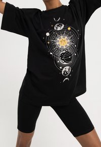 Person wearing black shorts and oversized black t-shirt with a celestial design featuring the sun, moon, and orbiting planets.