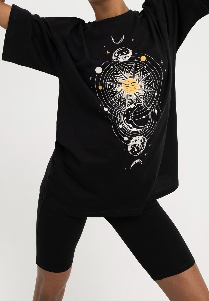 Person wearing black shorts and oversized black t-shirt with a celestial design featuring the sun, moon, and orbiting planets.