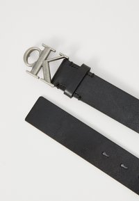 Ceinture en cuir noir avec texture embossée, dotée d'une boucle métallique argentée "CK". Comprend un passant en cuir et plusieurs trous de réglage.