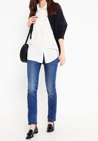 Černý kardigan s texturovanými rukávy přes bílou košili na knoflíky, doplněný modrými úzkými džíny a černými mokasínami.