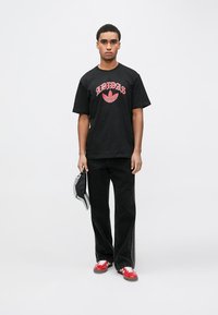 Černé tričko s červeným logem "Adidas", doplněné černými širokými kalhotami a červenými teniskami. Model drží šedou čepici.