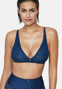 Femme posant en soutien-gorge en dentelle bleue et culotte taille haute assortie, se tenant devant un fond blanc avec une expression neutre.