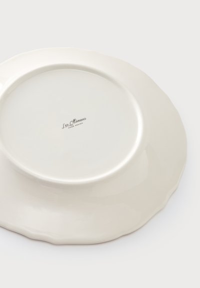 Assiette en céramique blanche à bords festonnés vue par le dessous, avec le logo "Les Ottomans Home Design" au centre.