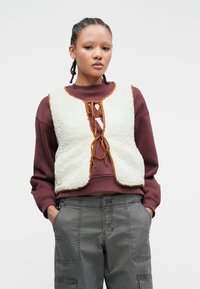 Neplederen vest in crèmekleur met bruine rand en vetersluiting, gedragen over een kastanjebruine sweatshirt; gecombineerd met grijze cargobroek.