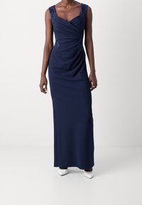 Navyblauwe maxi-jurk met kantaccenten op de schouders, draped voorkantontwerp en een gladde textuur. Gepaard met witte puntige schoenen.