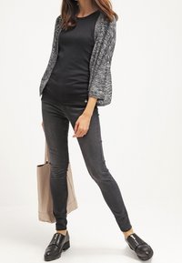 Mujer con camiseta negra, cárdigan gris abierto, vaqueros ajustados oscuros y zapatos negros sin cordones, sosteniendo una bolsa tote beige.