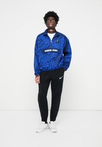 Blauwe met patronen Nike Air windbreaker jas met ritskraag en logo, gecombineerd met zwarte joggerbroeken en witte sportschoenen. Eenvoudig, sportief ontwerp.