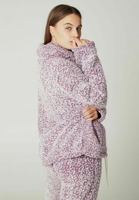 Violet pūkuotasis fliso megztinis su aukštu kaklu, tekstūruota paviršiumi ir laisvąja siluetu. Yra kengūrų kišenė ir reguliuojama raišteliai.