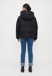 Mujer con cabello oscuro recogido en un moño, vistiendo una chaqueta acolchada negra con capucha, jeans azules con puños y botas negras de tacón, de espaldas sobre un fondo liso.