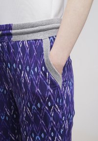 Lila mönstrade byxor med en diamantdesign och blå detaljer. Har grå elastisk midja och fickor. Slät tygtextur.