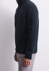 Pullover zip nero scuro realizzato in tessuto testurizzato, con colletto alto e silhouette aderente, abbinato a pantaloni grigi chiari.