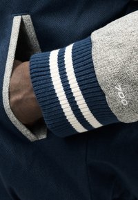 Χέρι που φοράει μανσέτα πλεκτή με μπλε και λευκές ρίγες, εν μέρει μέσα σε τσέπη παλτού σε σκούρο μπλε με γκρι υφασμάτινη άκρη και κεντητό λογότυπο «Y-3».