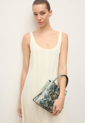 Mujer joven con un vestido blanco sin mangas sosteniendo un bolso de tamaño mediano con estampado de serpiente en tonos azul grisáceo y asa superior.