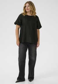 Schwarze strukturierte Bluse mit kurzen Puffärmeln, kombiniert mit dunklen geraden Jeans und spitzen schwarzen Schuhen, vor einem schlichten Hintergrund präsentiert.