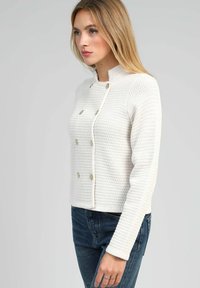 Veste blanche texturée avec col montant, design croisé et boutons dorés, associée à un jean bleu.