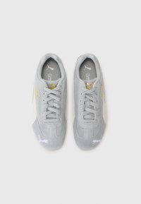 Baskets en daim gris avec des accents blancs, bout rond et lacets plats. Présente un logo doré sur le côté et une surface douce et texturée.