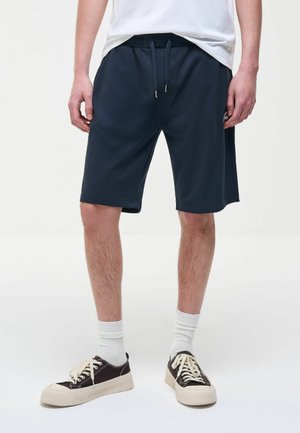 Uomo con pantaloncini blu navy con coulisse, calzini bianchi alla caviglia e scarpe da ginnastica nere basse con lacci bianchi, in piedi contro uno sfondo bianco.