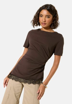 Braunes Trikot-Top mit kurzen Ärmeln, gerafften Seitendetails und Spitzenabschluss. Kombiniert mit hellbeigen Hosen. Glatte Textur.